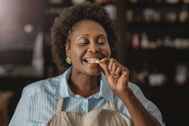 Smiling African American woman biting into a cookie at home Smiling African American woman standing with her eyes closed in her kitchen at home taking a bite from a cookie indulgence stock pictures, royalty-free photos & images