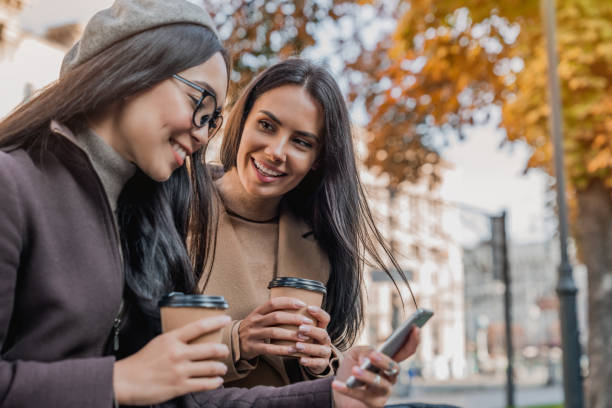 istock Two cheerful young girls using smartphone while sitting at the bench outdoors 1180000866
