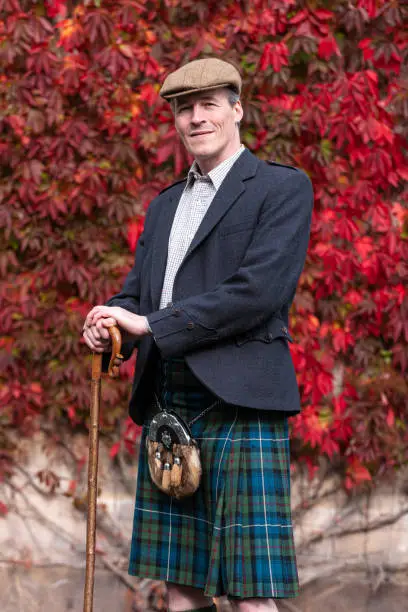 Elegant Scotchman in a traditional kilt and cloth cap posing with a walking stick or crook in front of a colourful red autumn creeper on a wall