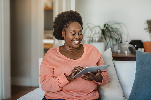 Smiling mature African American woman browsing the internet with a digital tablet while relaxing on her living room sofa at home