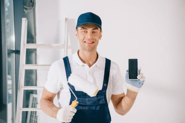 istock handsome young painter in uniform holding paint roller and showing smartphone with blank screen 1180038340