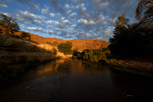 The sun is low, and highlight a cliff with warm light. The photo is taken with a wide angle lens which accentuates the river in the foreground, and the blue sky with clouds. The river is low. It is unusual because the river is flowing through a desert. The photo was taken close to Khowarib, near Sesfontein. Damaraland Namibia.
The photo was taken in February 2019.