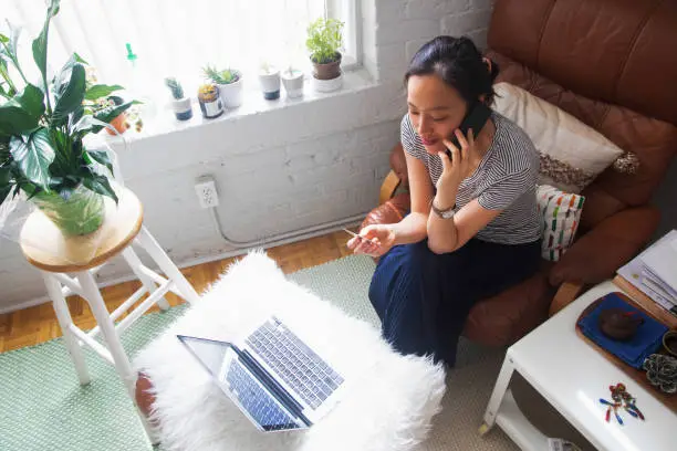 Overhead view of young Asian woman talking to customer service, credit card in hand, in her living room with a laptop in front of her.