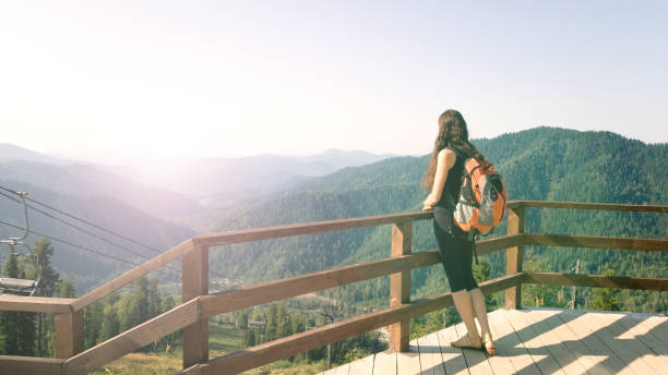 A brunette girl stands on an observation deck in the mountains and admires the beautiful view. A traveler with a backpack resting on the top of the mountain. Excursion to the Altai mountains. Dreamy and free concept stock photo