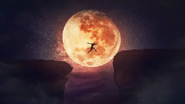 Photo of Surreal scene, self overcome concept, as determined man jump over a chasm obstacle. Way to win and success over starry night with full moon background. Motivation for achieving goals.