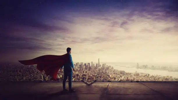 Photo of Rear view confident businessman, red cape suit, as hero stands on the rooftop of a skyscraper looking over the city horizon. Superhero leadership and success concept. Surreal super power metaphor.