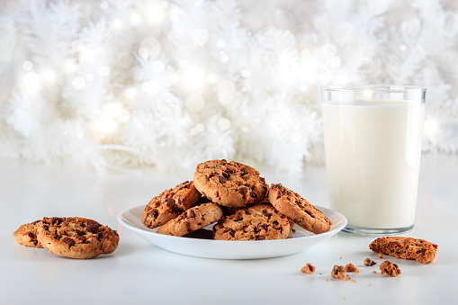 Delicious handmade chocolate chip cookies on a white plate and a glass of fresh milk on Christmas day. The lights are on decorating the white Christmas tree. Christmas holidays concept.