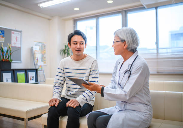 Male Japanese Patient Meeting with Senior Female Doctor Smiling young male Japanese patient meeting with senior female doctor in waiting room and going over results on digital tablet. japanese female doctor stock pictures, royalty-free photos & images