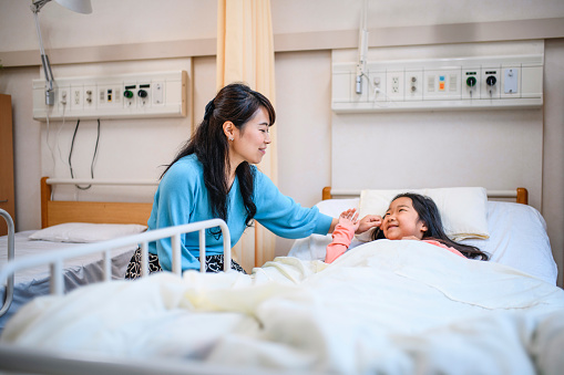 Smiling Japanese mother offering love and encouragement to young daughter lying in Tokyo hospital bed.