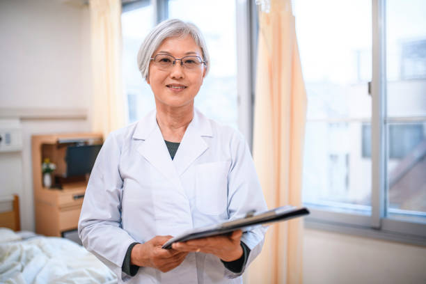 Relaxed Senior Female Japanese Doctor in Hospital Room Portrait of smiling senior female Japanese doctor standing with medical charts in Tokyo hospital room. japanese female doctor stock pictures, royalty-free photos & images