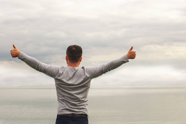 istock A young man is standing on the shore. The view from the back. Yoga classes. Hands raised up. Thumb up. Freedom and achievement. 1180106464