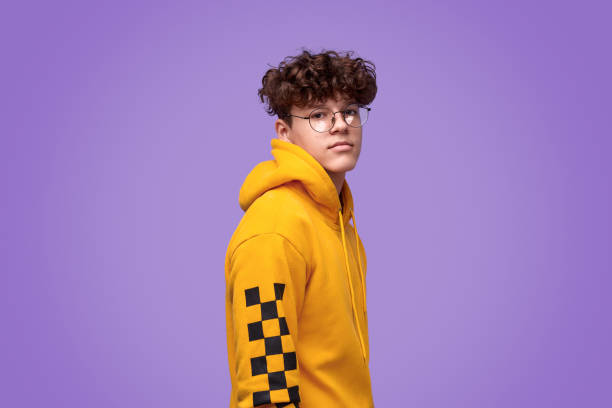 Stylish cool youngster looking at camera Side view of confident teenage in trendy yellow hoodie and round glasses looking at camera while standing against vivid purple background nerd teenager stock pictures, royalty-free photos & images