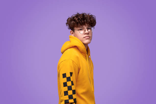 Stylish cool youngster looking at camera Side view of confident teenage in trendy yellow hoodie and round glasses looking at camera while standing against vivid purple background hipster person stock pictures, royalty-free photos & images