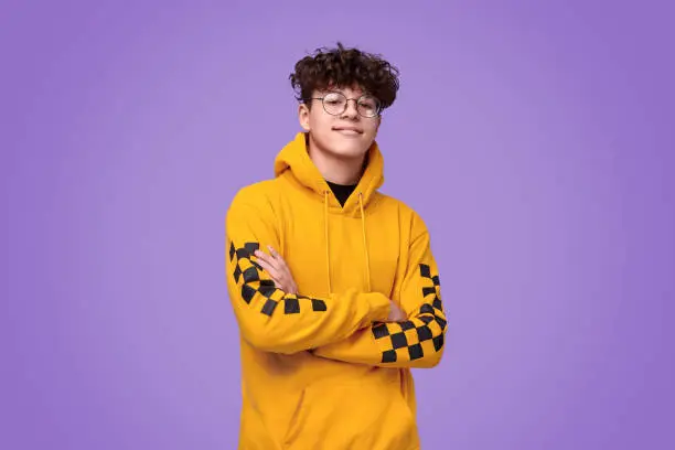 Positive teen boy in yellow hoodie crossing arms and looking at camera against bright violet background