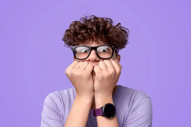 Shocked youngster in glasses biting nails and looking at camera in fear against bright purple background