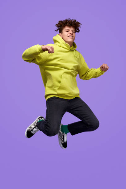 Stylish teenager jumping up in air Full body happy youngster in trendy outfit looking at camera and jumping high against vivid purple background jumper stock pictures, royalty-free photos & images