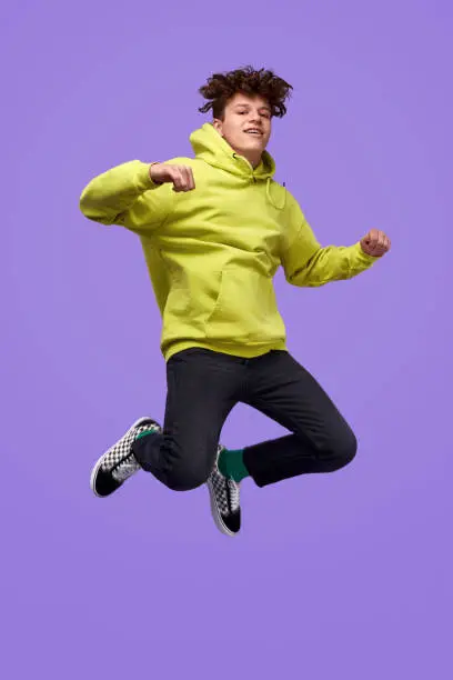 Full body happy youngster in trendy outfit looking at camera and jumping high against vivid purple background