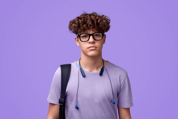 Teen student looking seriously at camera Nerdy youngster with backpack and earphones looking at camera while standing against purple background blank expression photos stock pictures, royalty-free photos & images