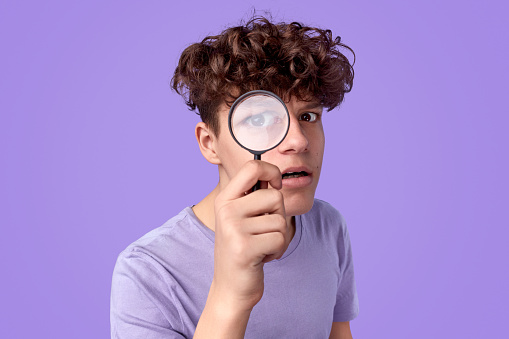 Curious teen boy with looking at camera through magnifying glass while searching for clues against violet background