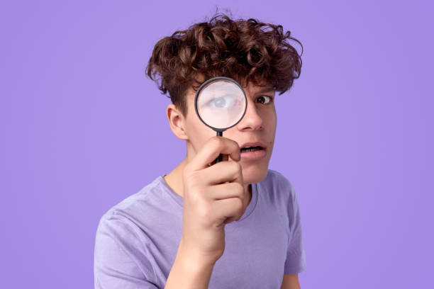 Young detective looking at camera Curious teen boy with looking at camera through magnifying glass while searching for clues against violet background curiosity stock pictures, royalty-free photos & images