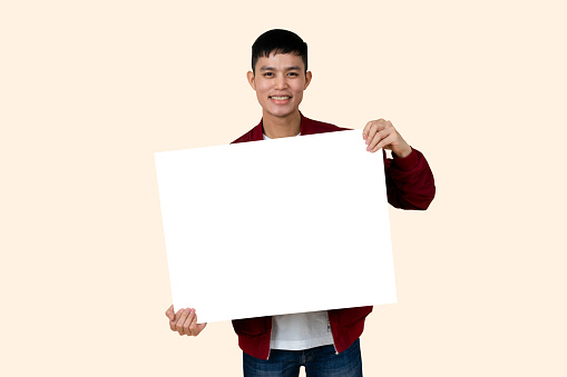 close up young asian ethnicity teenage man showing mockup blank poster (for add content) and smiling with happiness feeling isolated on cream color background for advertisement banner and billboard design concept