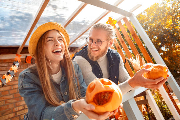 ハロウィーンプレパラトンコンセプト。彫刻されたカボチャで家を飾るフェンスに寄りかかっている若いカップルは、陽気な側面のクローズアップを笑う - photography close up side view halloween ストックフォトと画像