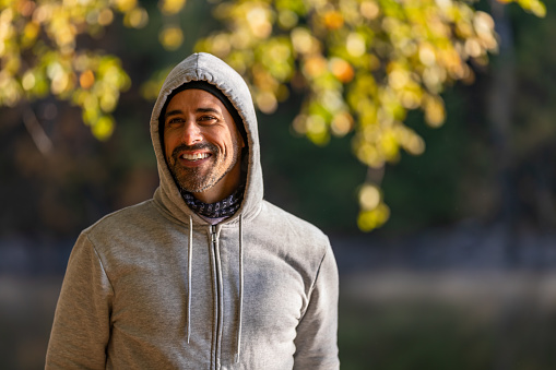 A Latino man wearing a grey hoodie is smiling. He is standing in a forest.