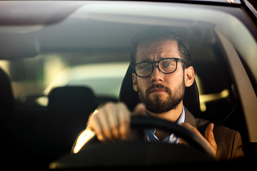 Photo of caucasian serious bearded businessman in formal wear driving car during the day.