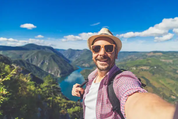 Cheerful man traveler with straw hat and sunglasses taking selfie against beautiful valley. Travel and nature explore concept. Space for copy.