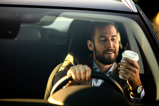 Photo Of happy Business Man Driving Car. Successful Young Smiling Businessman Drinking Coffee From Paper Cup While Driving Car. Handsome Happy Male Going To Work In Comfortable Car. High Resolution.
