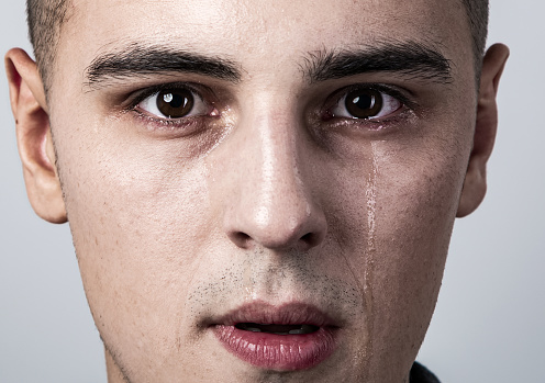 Unhappy crying young man with dropping tears on the face. Closeup studio portrait on grey background. Negative emotional concept. Toned dark shadow color