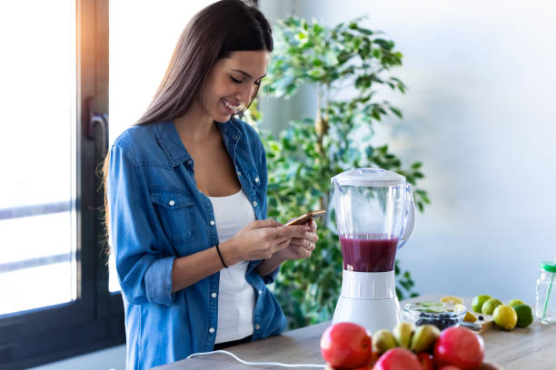 pretty young woman using her mobile phone while preparing fruit smoothie in the kitchen at home. - blueberry fruit berry berry fruit imagens e fotografias de stock
