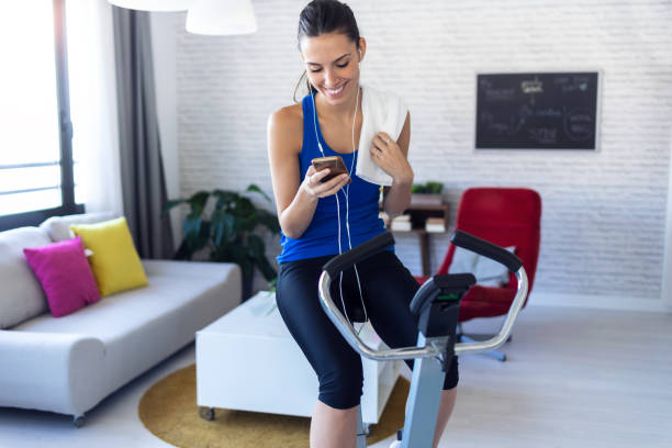 Smiling fitness girl using mobile phone after training on exercise bike at home. Shot of smiling fitness girl using mobile phone after training on exercise bike at home. exercise bike stock pictures, royalty-free photos & images