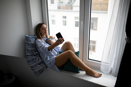 Young woman relaxing on window sill and taking selfie