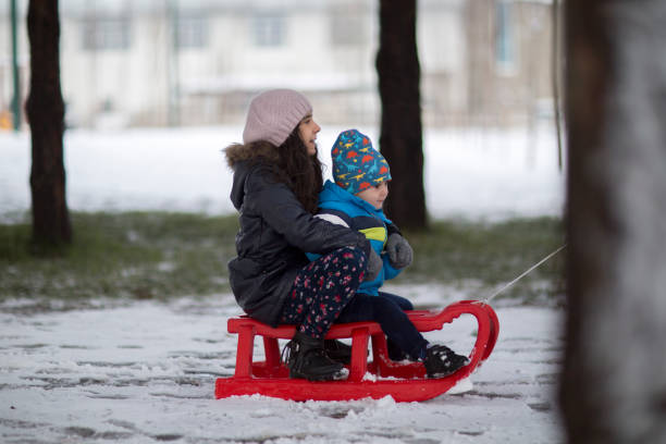A mother helping her young child ride a sled in a snowy park, outdoor winter activity, parenting, childcare