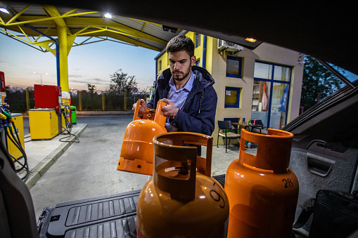 Young man getting out of car trunk orange lpg tanks with gas at night , in front of gas station