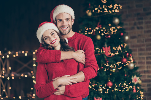 Photo of cute couple spending christmas time eve in decorated garland, lights room standing piggyback near x-mas tree indoors wear red pullovers and santa hats