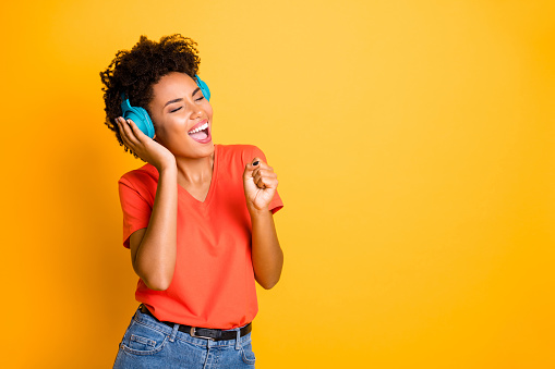 Copyspace photo of curly wavy cheerful charming lady singing into her imaginary, microphone wearing jeans denim wireless earphones isolated over yellow vibrant color background