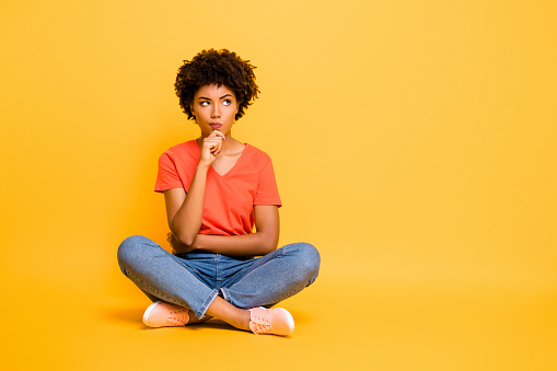 Full length body size photo of pensive solving deciding minded girlfriend, sitting with her legs crossed wearing jeans denim t-shirt touching her chin isolated over yellow vivid color background