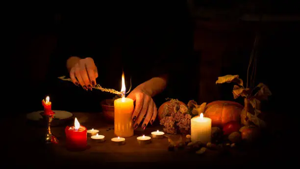 Witch make a spell on the altar in the dark. Female hands with sharp black nails burning magic herb among candles, pumpkin, nuts, dry leaves, selected focus, low key. Halloween, Yule, witchcraft, voodoo, fortune telling and divination concept