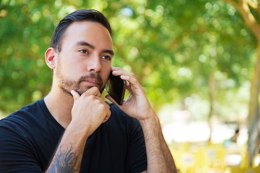 Pensive guy talking on cellphone outdoors. Handsome man with tattooed arm speaking on mobile phone on park, touching chin, looking away. Phone talk outside concept