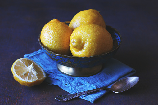 Low key dark and moody photo of a bright yellow juicy lemons in a blue antique bowl standing on a blue napkin with a spoon taken in the natural light