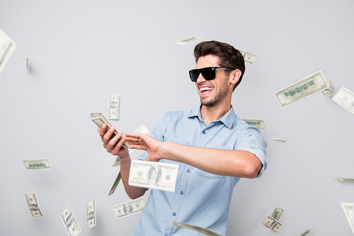 Photo of cheerful excited ecstatic overjoyed man throwing money away, showing his wealthiness wearing denim isolated over grey color background