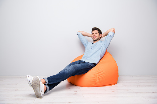 Turned full length body size photo of cheerful confident, relaxing man sitting in chair thinking guessing smiling toothily having rest fun isolated over grey color background