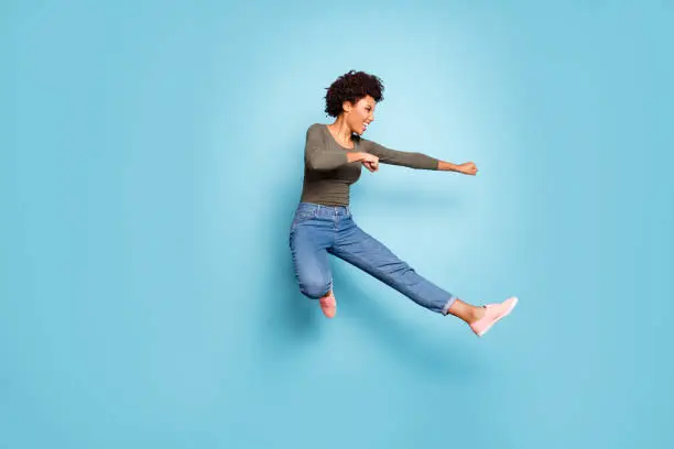 Full length photo of dark skin wavy lady jumping high dancing modern, youngster moves practicing judo fighting wear casual outfit isolated pastel blue color background