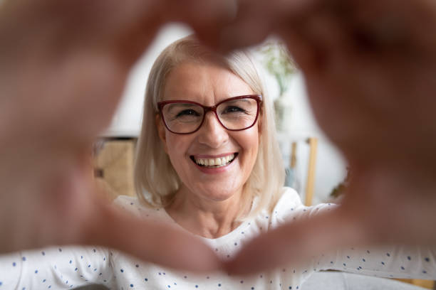 mulher madura de sorriso do retrato que olha através das mãos dadas forma coração - eyesight senior adult care support - fotografias e filmes do acervo