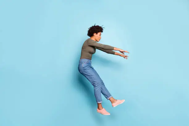 Photo of Oh my god. Full length photo of dark skin wavy lady jumping high blown away by strong stormy wind confused wear casual outfit isolated pastel blue color background