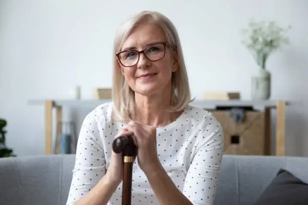 Head shot portrait beautiful older woman in glasses with wooden cane sitting on couch at home, mature female holding hands on walking stick, looking at camera, older people healthcare concept