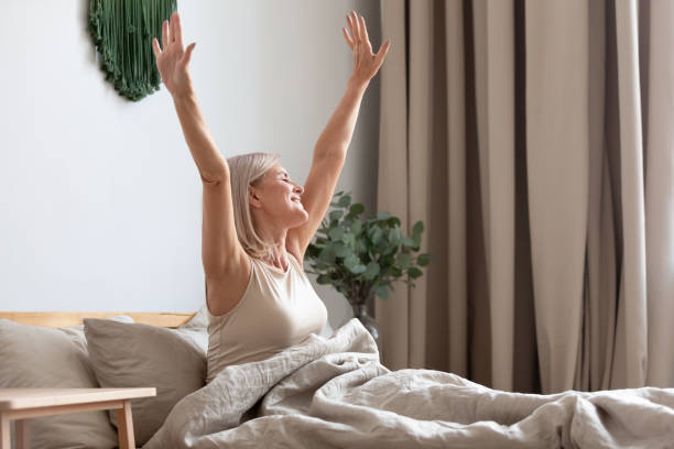 ベッドに座って幸せな年配の女性、目覚めた後に手を伸ばす - bed cheerful enjoyment excitement ストックフォトと画像