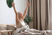 Happy older woman sitting in bed, stretching hands after awakening